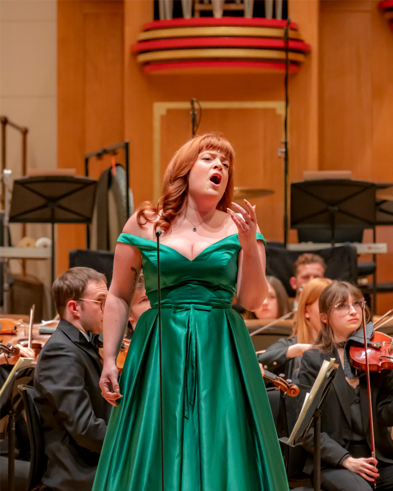 A student performing on stage in a green dress with an orchestra behind her.